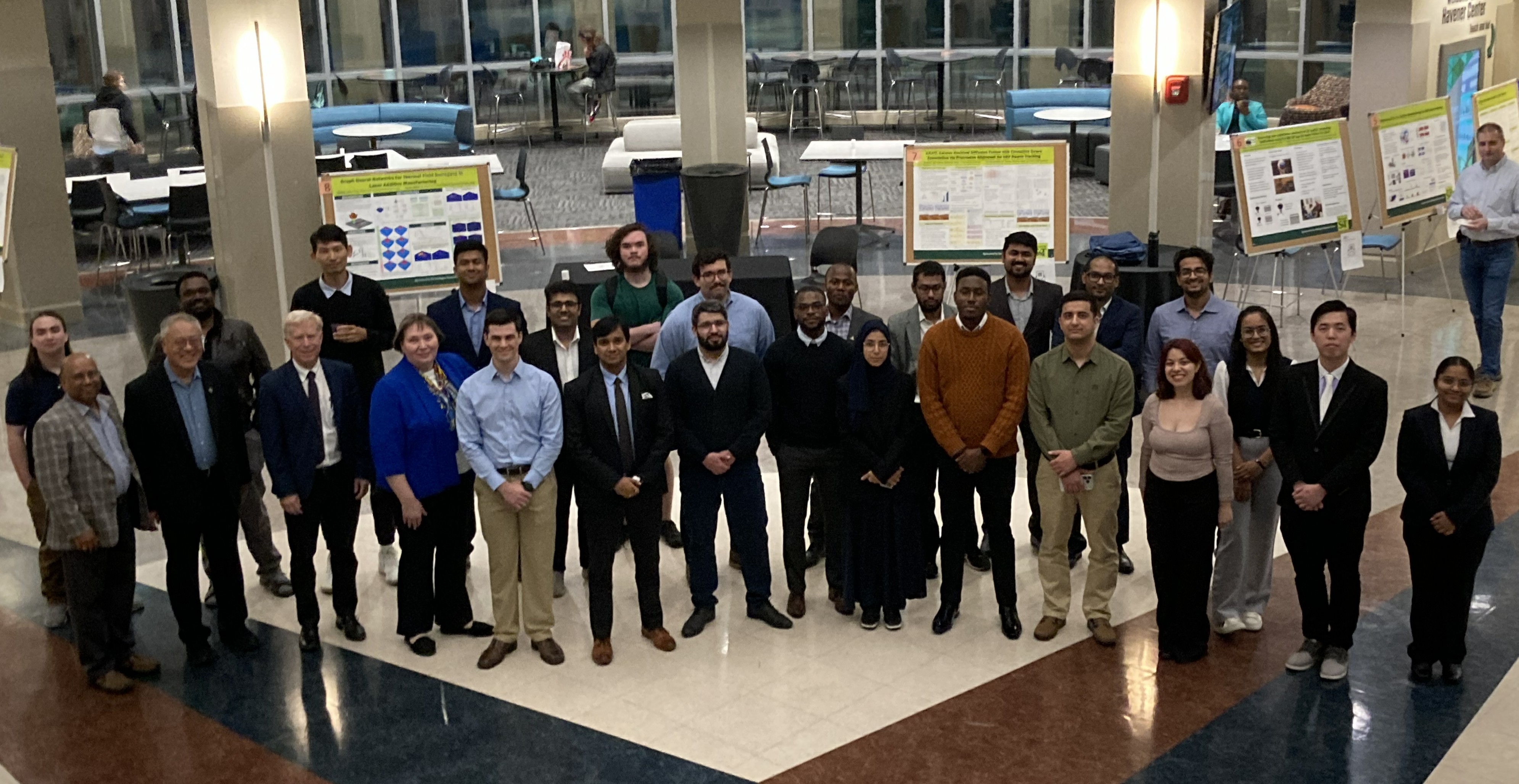 A diverse group of 27 people poses for a photo in a spacious hall, with research posters displayed in the background, conveying an academic atmosphere.